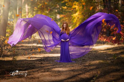 Sesja ciążowa: kobieta w ciąży, ubrana w rozwianą fioletową sukienkę, stoi w lesie.
