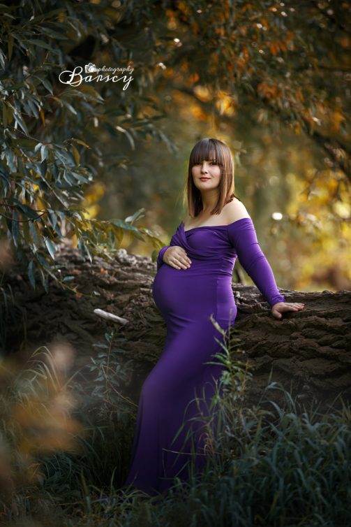 Kobieta w ciąży w fioletowej sukience pozuje oparta o drzewo podczas sesji ciążowej.