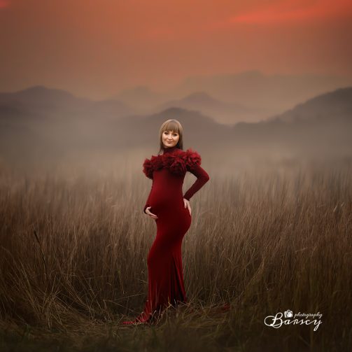 Kobieta w ciąży w czerwonej sukience stojąca w polu na tle gór.