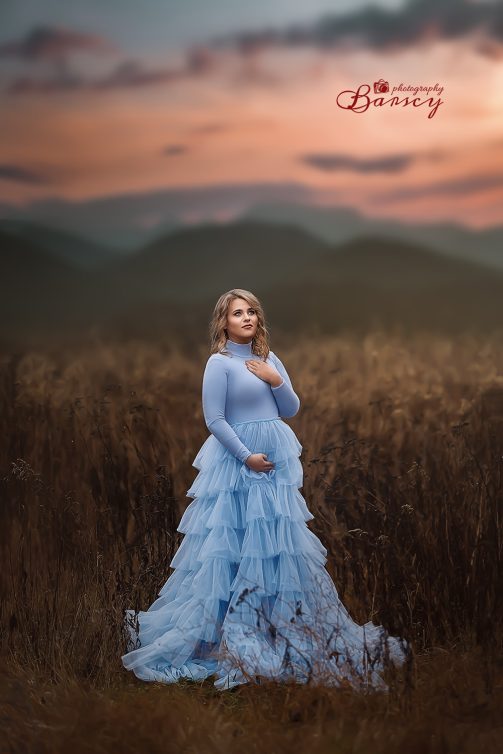 Kobieta w ciąży w niebieskiej sukience stojąca na polu o zachodzie słońca.